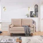 Goldoma 57-inch beige velvet loveseat with tufted seat, track arms, dark brown wood legs, and two toss pillows in a modern living room