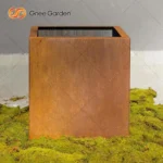 Large corten steel planter filled with tall grasses, set against a brick wall in a garden setting.