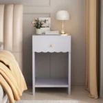 Modern nightstand with scalloped edge drawer and open shelf in lilac color, placed beside a bed