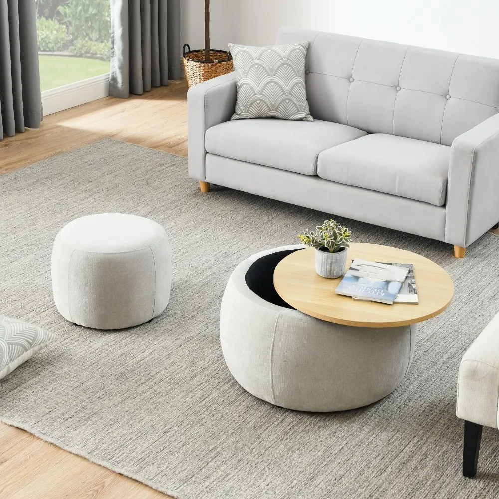 H22236f6aebc34b17a4cb0403ba8bd2d6I.webp Goldoma round storage ottoman in beige fabric with hinged lid, used as end table with a cup on top, in a living room setting