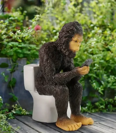Whimsical resin sasquatch garden statue sitting on a toilet, brown and gray textured finish, placed on a lush green