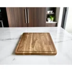 Brown square solid wood cutting board with juice groove and utensils on kitchen counter