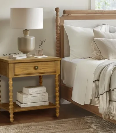 Natural wood nightstand with one drawer and a shelf beside a bed in a bedroom