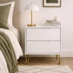White nightstand with gold hairpin legs featuring two storage drawers in a modern bedroom setting