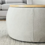 Goldoma round storage ottoman in beige fabric with hinged lid, used as end table with a cup on top, in a living room setting