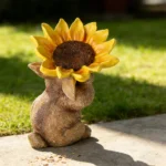 Resin rabbit with sunflower hat among flowering plants, outdoor yard decoration.
