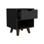 Modern nightstand with drawer and open shelf in black finish with wooden legs on a bedroom floor next to a bed