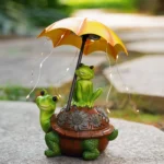 Frog on turtle statue illuminated at night, warm LED glow, umbrella casts light pattern, garden pathway setting.