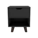 Modern nightstand with drawer and open shelf in black finish with wooden legs on a bedroom floor next to a bed