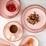 Elegant dinnerware set with gilt rim pink porcelain plates, bowls, mugs, and cutlery on a table