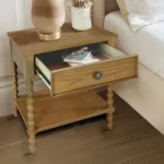 Natural wood nightstand with one drawer and a shelf beside a bed in a bedroom