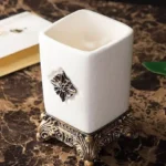 Mouthwash cup with metallic gold rim, sitting next to a matching soap dish on a wooden bathroom shelf.