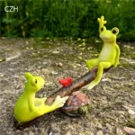 Frog playground ornament, seesaw with two frogs, one green with yellow belly, other brown with spots.