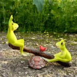 Side view of cute frog couple balancing on seesaw, colorful painted details, surrounded by small stones.
