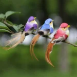 Three artificial birds in red, yellow, and green placed among potted succulents on a patio table, outdoor setting.
