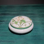 Ceramic lotion bottle with flower design, on bathroom shelf with plants.