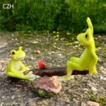Frog couple figurine on seesaw, one frog's arm raised, other's leg up, playful action, on moss.