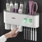 Automatic toothpaste dispenser in white, mounted on bathroom wall with toothbrush holder and cup set.