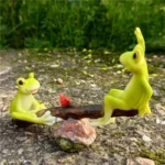 Frog seesaw figurine with one frog lifting off, playful pose, in a fairy garden with tiny flowers.