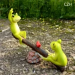 Cute frog couple on seesaw, one frog wearing a tiny hat, other with a scarf, in outdoor setting.