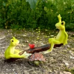 Cute seesaw frog miniature figurine couple on wooden seesaw, green and brown, in fairy garden setting with pebbles.