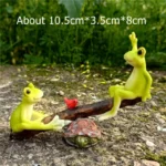 Miniature frog seesaw with one frog holding a tiny flower, in a fairy garden with white pebbles.