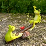 Close-up of frog couple figurine, one frog with red hat, the other with bow tie, on miniature seesaw.
