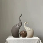Nordic matte ceramic elephant family figurines on a modern white bookshelf, soft natural lighting.