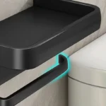 Toilet paper holder in bathroom corner, with shelf holding extra rolls and accessories.