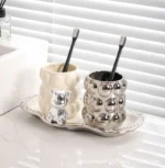 Pair of electroplated ceramic cups on a bathroom shelf with towels, one with a toothbrush and the other empty.